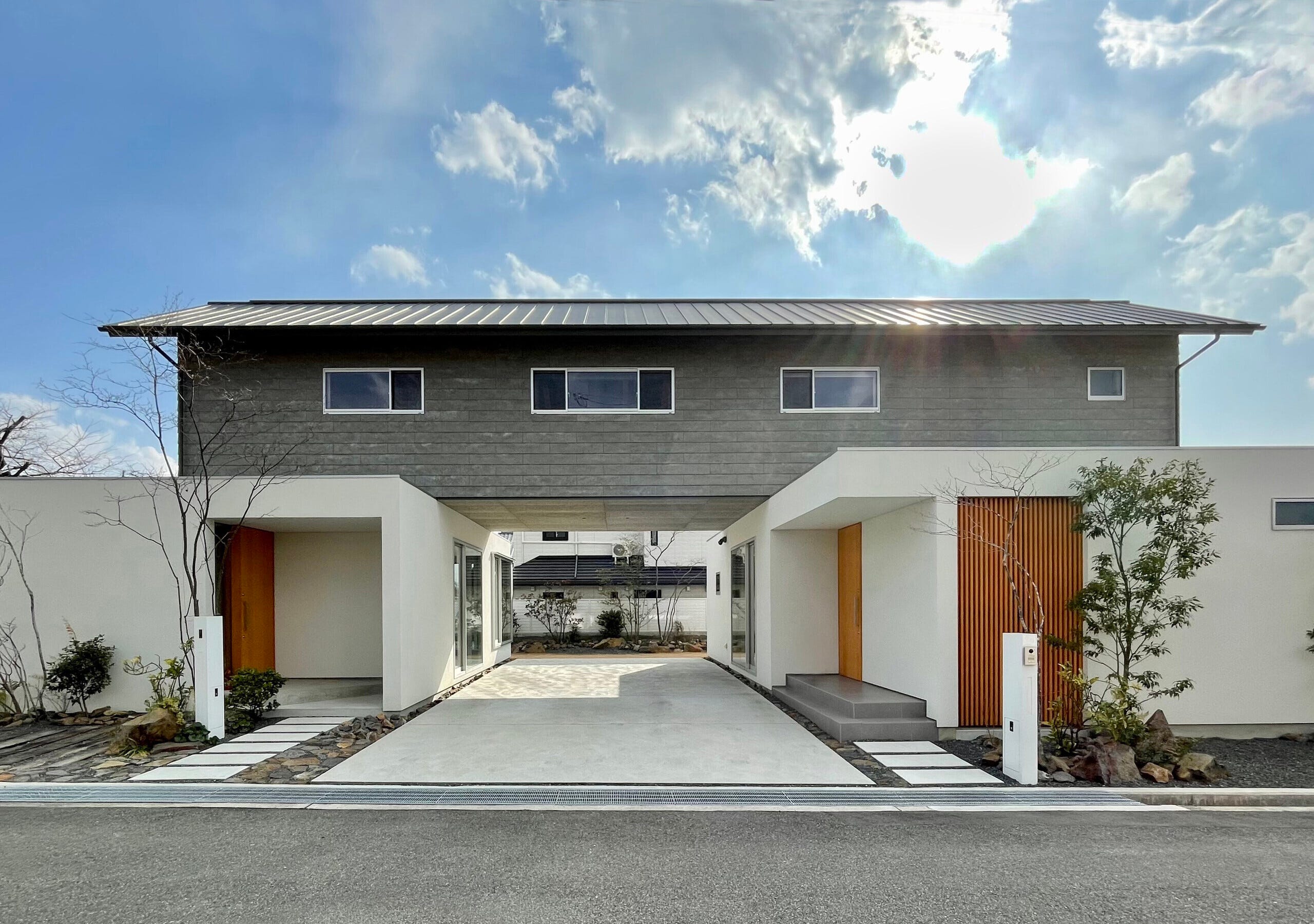 施主からは完全分離型の二世帯住宅が求められるなか、必然的に大きくなりがちな建物ボリュームを抑えながら、両世帯の距離感をどう設計するかをテーマに、二世帯住宅の在り方を模索しました。また、郊外に暮らす家族にとって必要な車の台数も多く、駐車スペースの在り方についても検討が必要でした。
そこで、LDKからなる1階ヴォリュームを共有の通り庭を挟んで両世帯それぞれ独立配置し、その上に積層するように2階ヴォリュームを架け渡しました。そこに生まれたトンネル状に抜ける軒下空間は普段駐車スペースとして利用されており、互いに距離を保つバッファゾーンになっていますが、車を前に動かせば両世帯を繋ぐテラスとなり夏のBBQや子供たちのプール遊びなど、さまざまなライフシーンを彩ります。使い方次第で繋いだり隔てたりと距離感を変化させ、お互いの様子を感じつつも両世帯のプライバシーを確保できるように配慮されています。
また、架け渡された2階は周辺住宅に馴染むよう切妻屋根とし、道路からセットバックした位置に配置することで、周囲の桜並木通りの景観にも配慮しています。その2階は、主にそれぞれの寝室からなるプライベートな空間です。中央にファミリールームがあり、両世帯を内部で行き来できる唯一の場所でもあります。将来は子供室として分割することで、ライフスタイルの変化にも対応できるように計画してあります。更には、将来の世帯数の変化に伴い、片方の住戸を賃貸として貸すことも考えられます。家族構成が変わるたびに建て替えるのではなく、様々な住まいの在り方への変化を許容できる、そんな住宅になったのではないでしょうか。